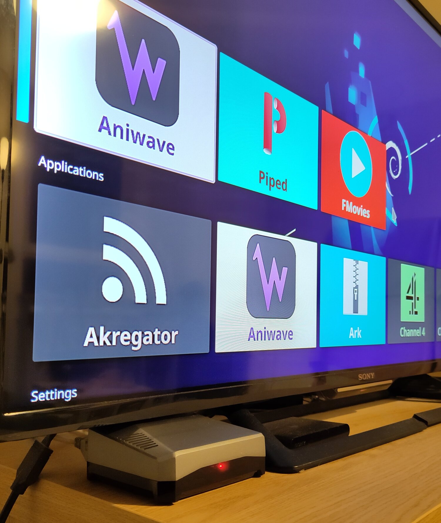 A Rspberry Pi 4b in an Argon One M.2. Sata case, powered
        on and plugged into a TV that is on the KDE Plasma Bigscreen
        home screen, with a Debian logo wallpaper.