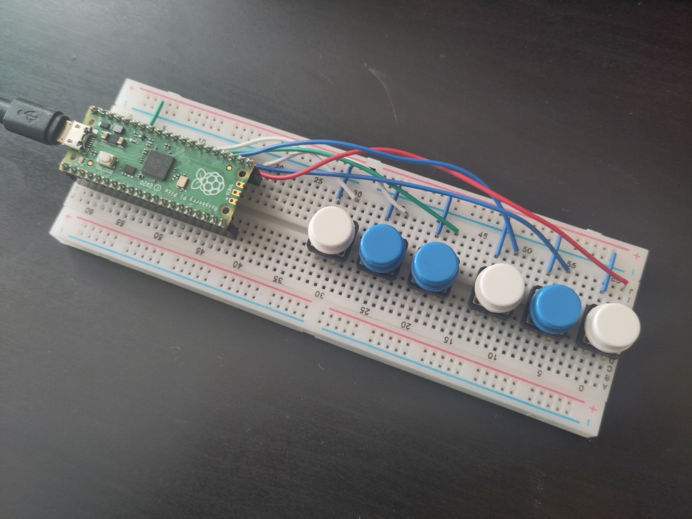 A Raspberry Pi Pico controller on a breadboard, wired
        up to six coloured buttons, in the pattern of white, blue,
        blue, white, blue, white.