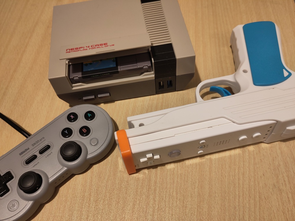 A NesPi-4 cased Raspberry Pi 4b, with the slot open
        to show a NES cartridge style SSD case, an 8BitDo SN30 Pro
        controller, and a WiiMote in a pistol lightgun adapter.