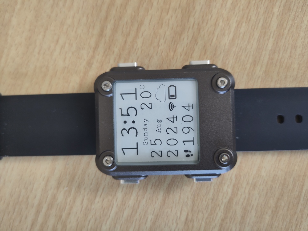 A Watchy with gun metal case displaying the time, date,
        weather and a steps count in a custom monospace font.